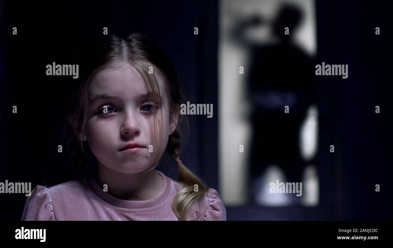 Crying little girl looking at camera silhouette of alcoholic father outside door Stock Photo