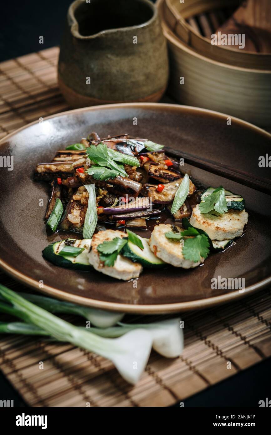 Spicy Aubergine / Eggplant with tofu and vegetable stir fry, served on a ceramic plate with chopsticks. Bamboo matt and steamer with black background Stock Photo