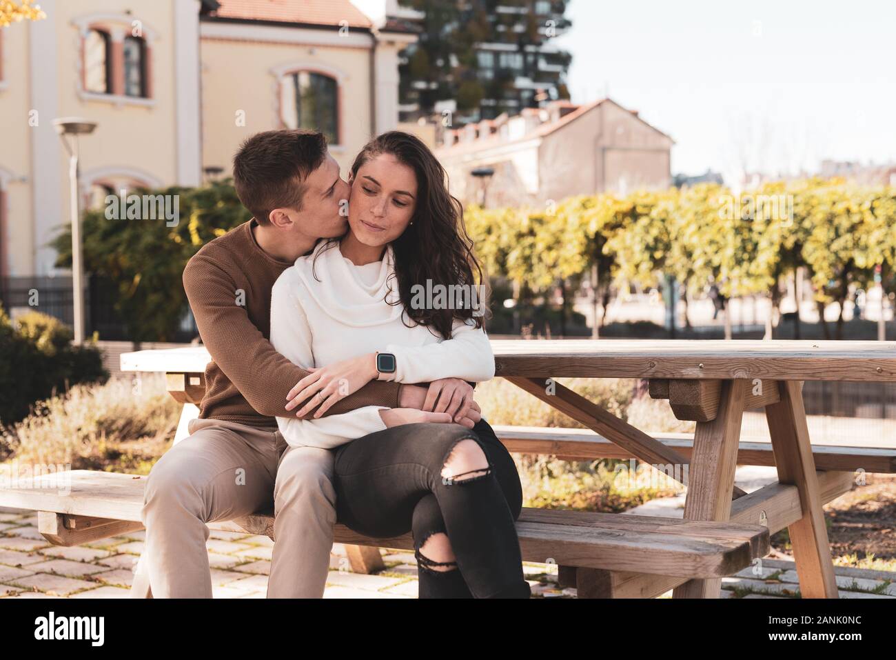young couple of millenials in love hugging on a bench, he kisses her gently, she is happy, love concept Stock Photo
