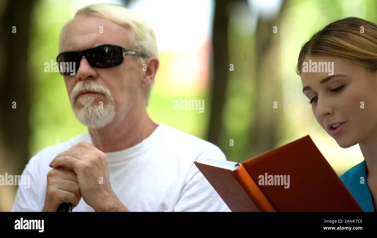 Nurse reading book to old blind man, resting outdoor near clinic for