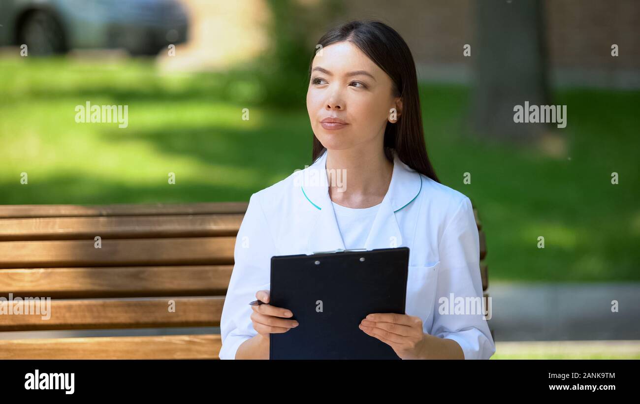 Thoughtful female doctor working outside, sitting on bench in hospital park Stock Photo