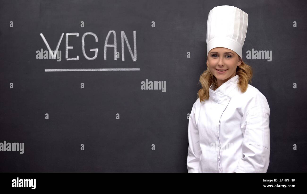 Smiling chief cook standing against black board with vegan word, dietology Stock Photo