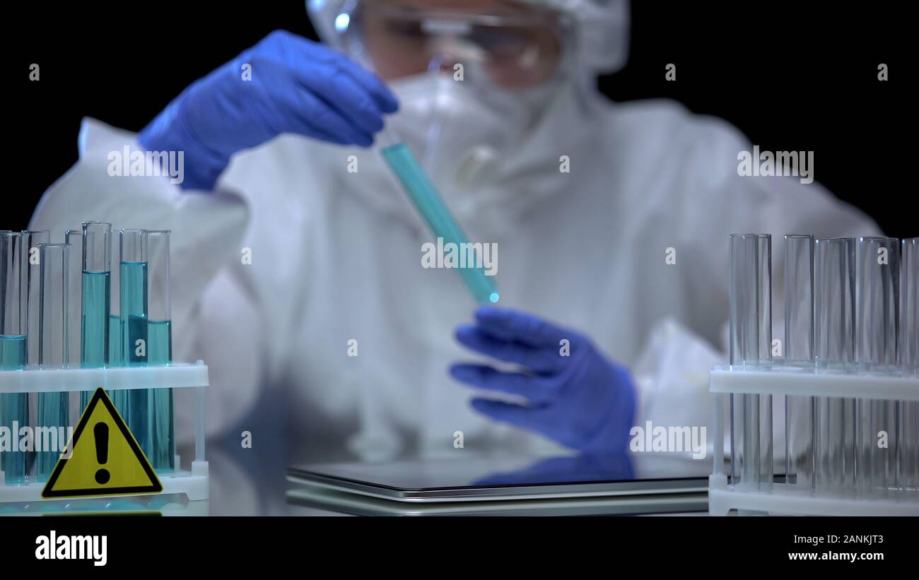 Scientist in protective suit analyzing dangerous liquid tubes, tablet on table Stock Photo