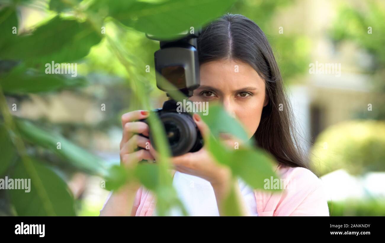 Suspicious female with camera hiding behind trees at park, spying husband Stock Photo