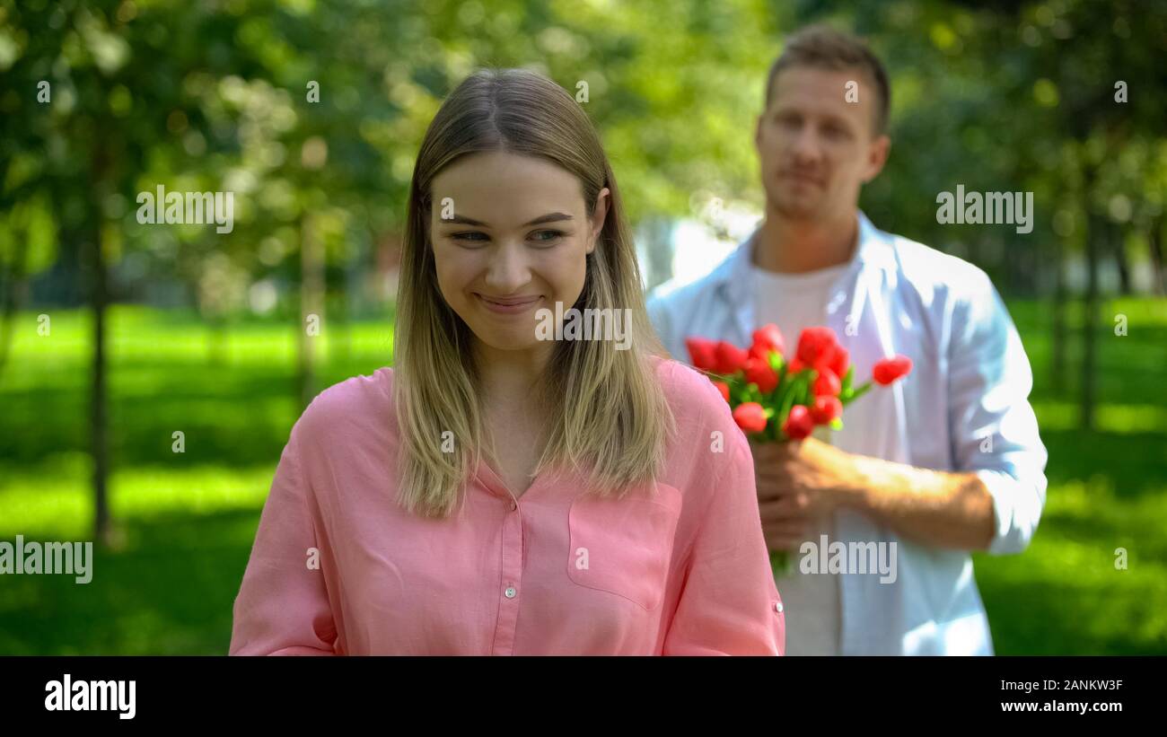 Happy smiling woman in park, boyfriend with flowers behind, date invitation Stock Photo