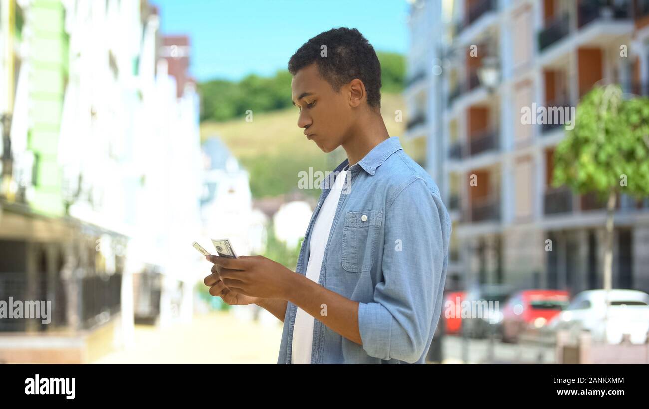 Upset mixed-race teen boy counting dollars near shop sad about purchase sum lack Stock Photo