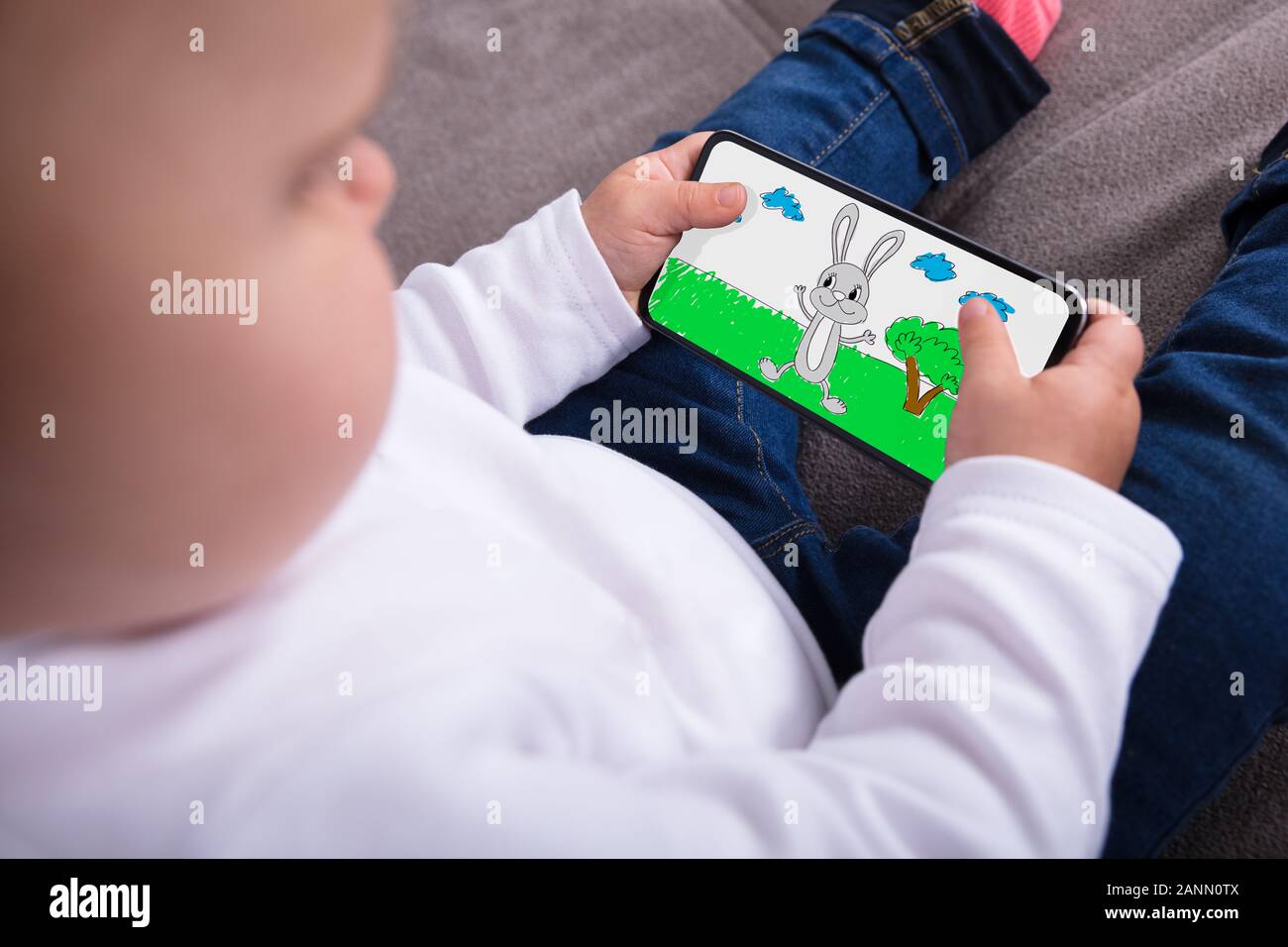 Happy Baby Girl Sitting On Sofa Watching Cartoon On Smartphone Stock Photo