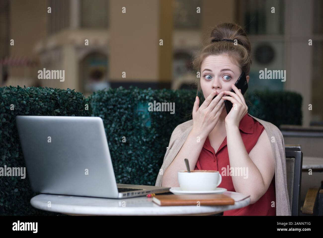 Young caucasian woman in cafe with coffee and laptop, talking on the phone with shocked, surprised expression Stock Photo