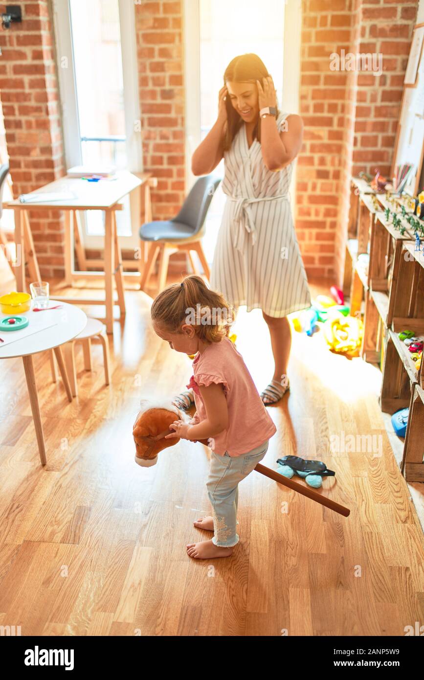 Beautiful teacher and blond student toddler girl ridding horse toy with stick at kindergarten Stock Photo