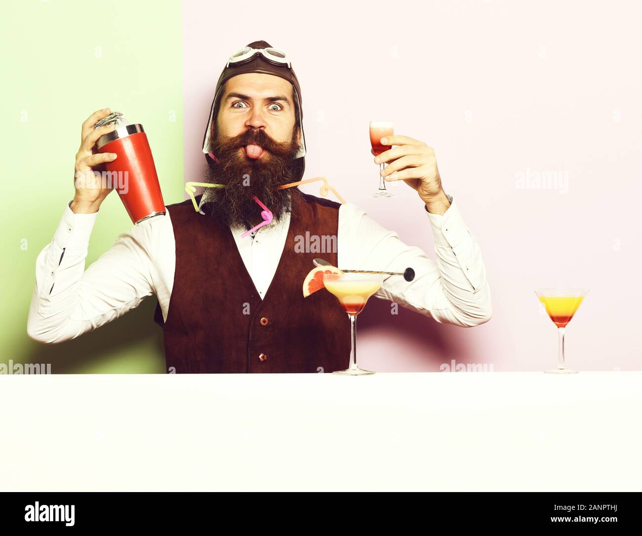 handsome bearded pilot with long beard and mustache on funny face with shaker alcoholic cocktail in vintage suede leather waistcoat with hand ,and glasses on purple green studio background Stock Photo