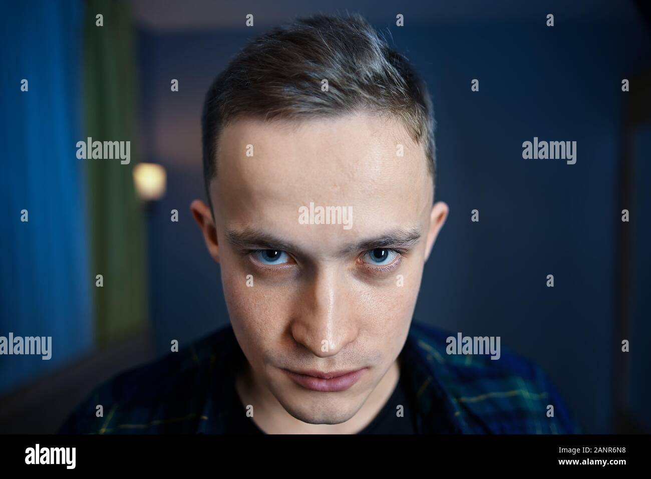 Young man with paranoid schizophrenia looks out from under his eyebrows with hatred. An embittered guy with gray eyes and a short haircut against a da Stock Photo