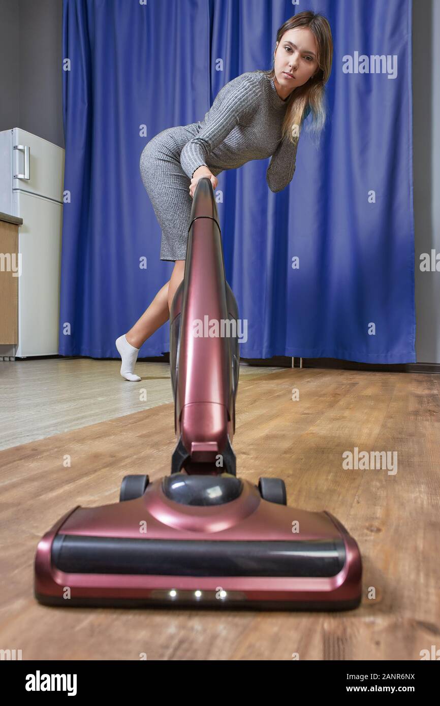 Thin housemaid uses a stick vacuum cleaner when cleaning floor from dust. A young woman in a tight-fitting dress is vacuuming room with an electric br Stock Photo