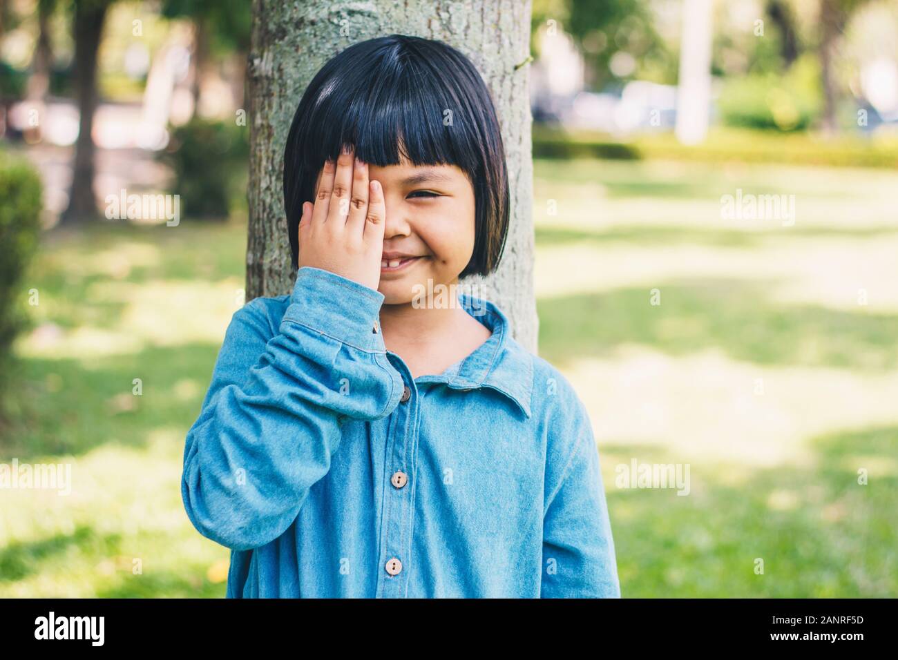 The image of a little girl with her hands closed, her right eye leaning against a tree in a park. Stock Photo