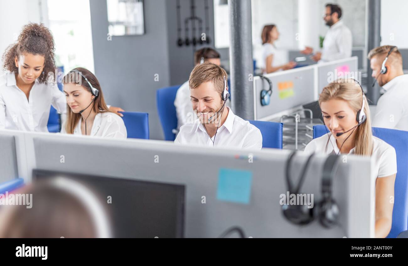 Call center worker accompanied by his team. Smiling customer support operator at work. Young employee working with a headset. Stock Photo