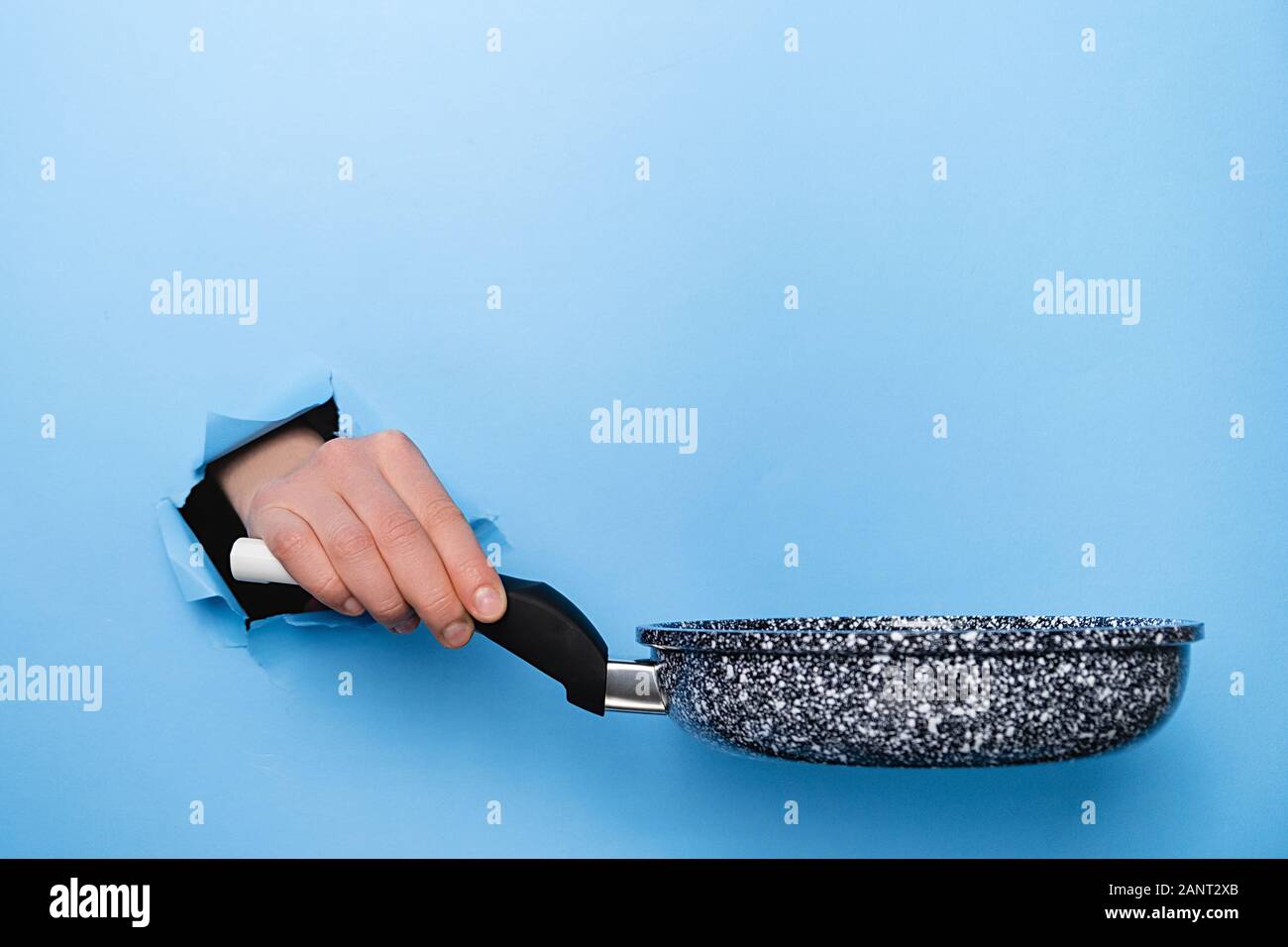 Close up of female hand holding Frying pan through torn paper blue background. Kitchen utensils concept. Copy space for advertisement Stock Photo