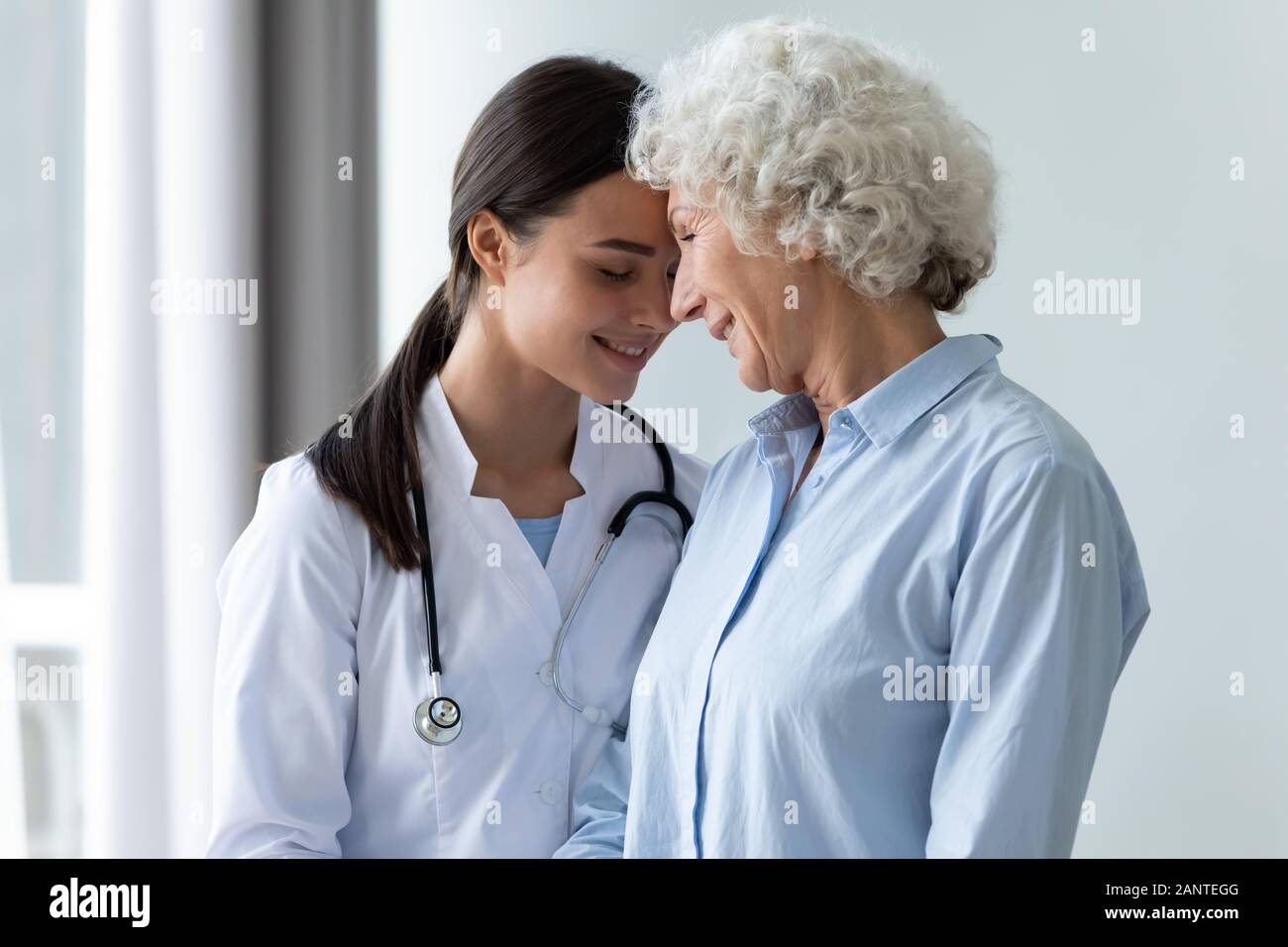 Smiling girl caregiver and happy older woman touching foreheads Stock Photo