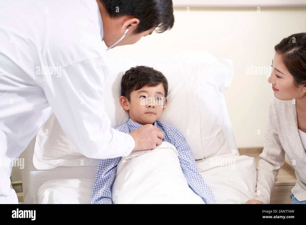 five year old asian kid lying in bed in hospital ward accompanied by mother and being examined by pediatrician using stethoscope Stock Photo