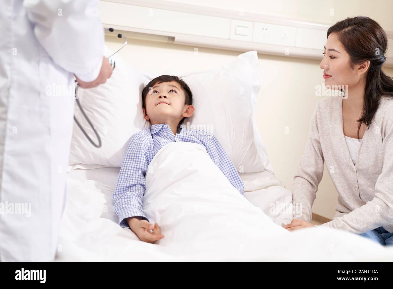 five year old asian child lying in bed in hospital ward accompanied by mother talking to doctor Stock Photo