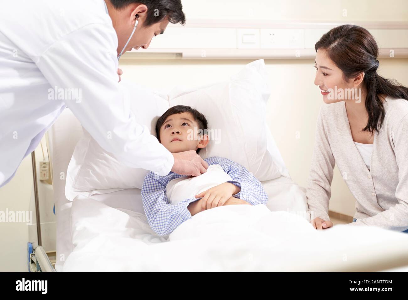five year old asian kid lying in bed in hospital ward accompanied by mother and being examined by pediatrician using stethoscope Stock Photo