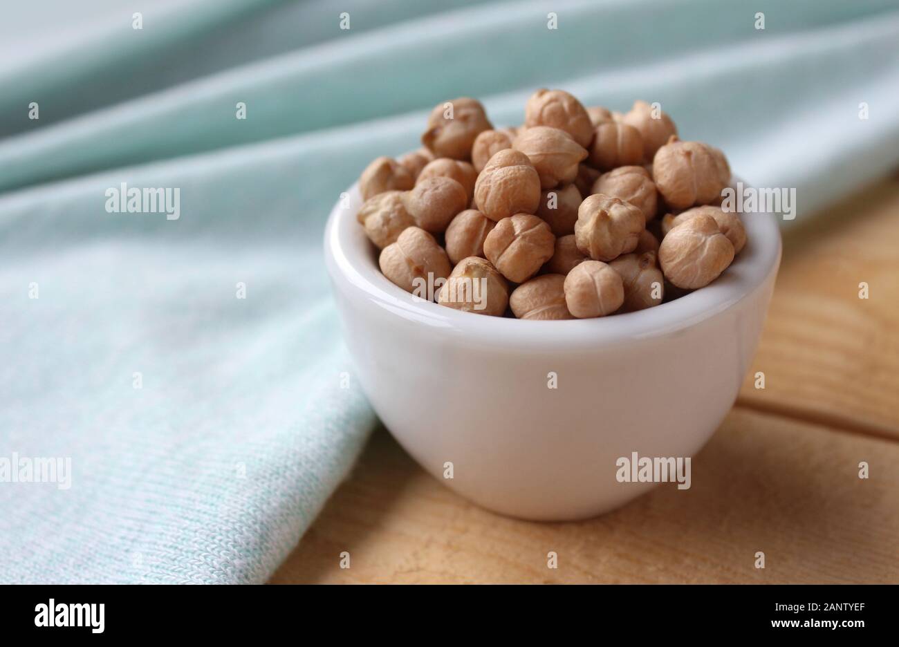 Side on view of a white bowl filled with chick peas (Cicer arietinum), on a wooden table with mint green table cloth. With copyspace. Stock Photo