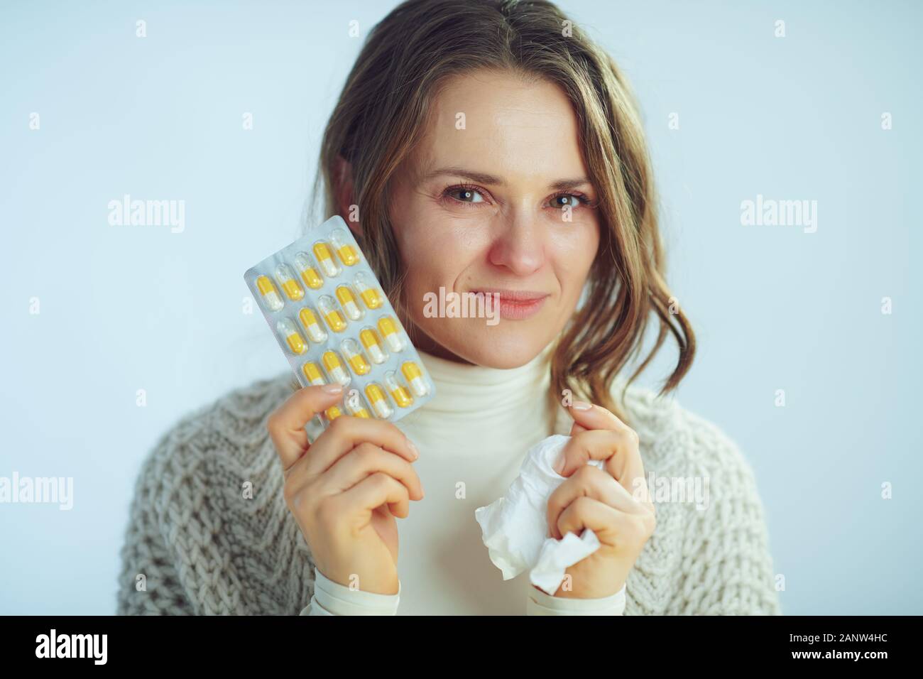 Portrait of smiling ill modern woman in roll neck sweater and cardigan with napkin showing blister pack of pills on winter light blue background. Stock Photo