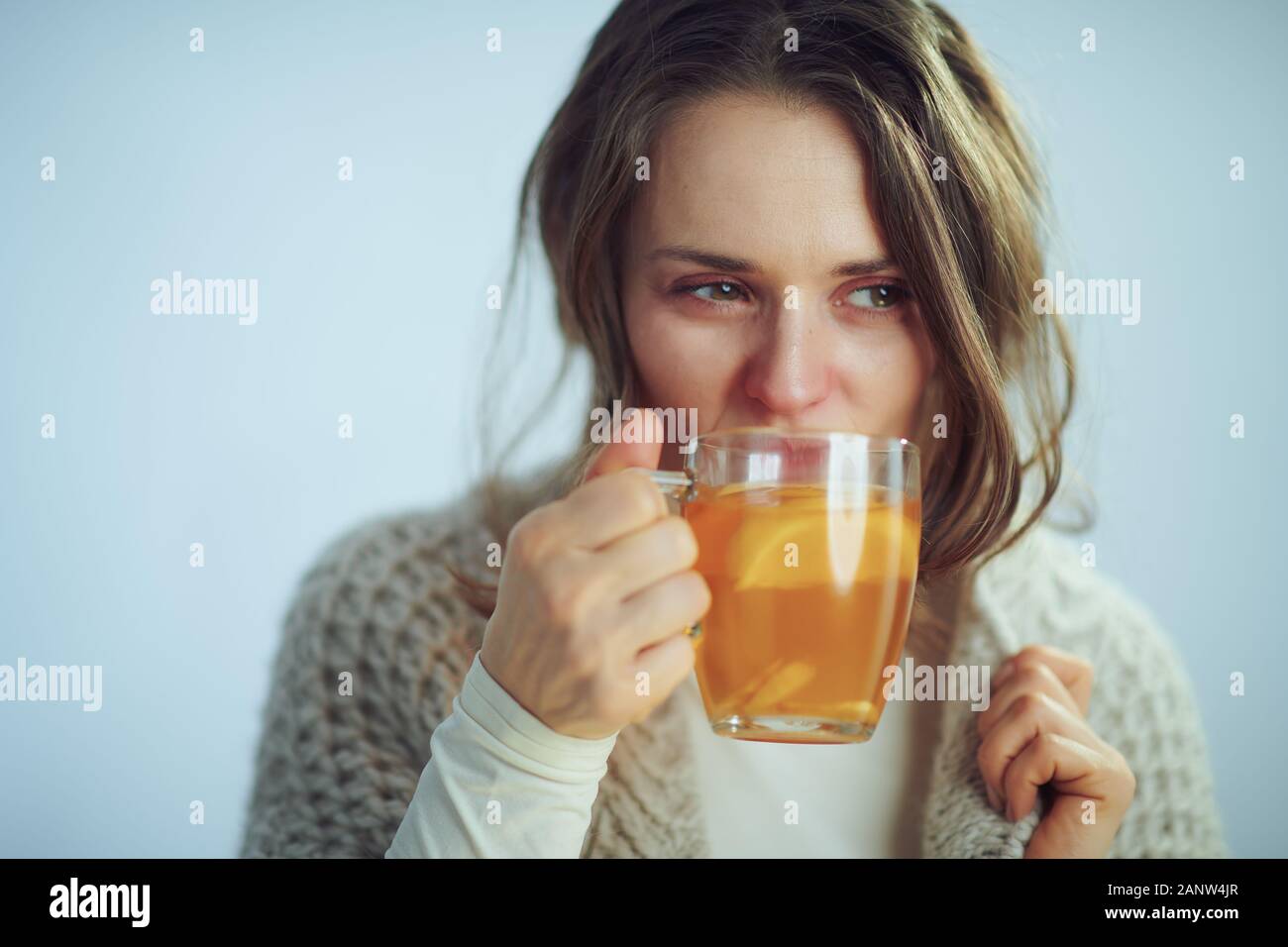 ill elegant housewife in roll neck sweater and cardigan drinking cup of hot tea with ginger, lemon and honey on winter light blue background. Stock Photo