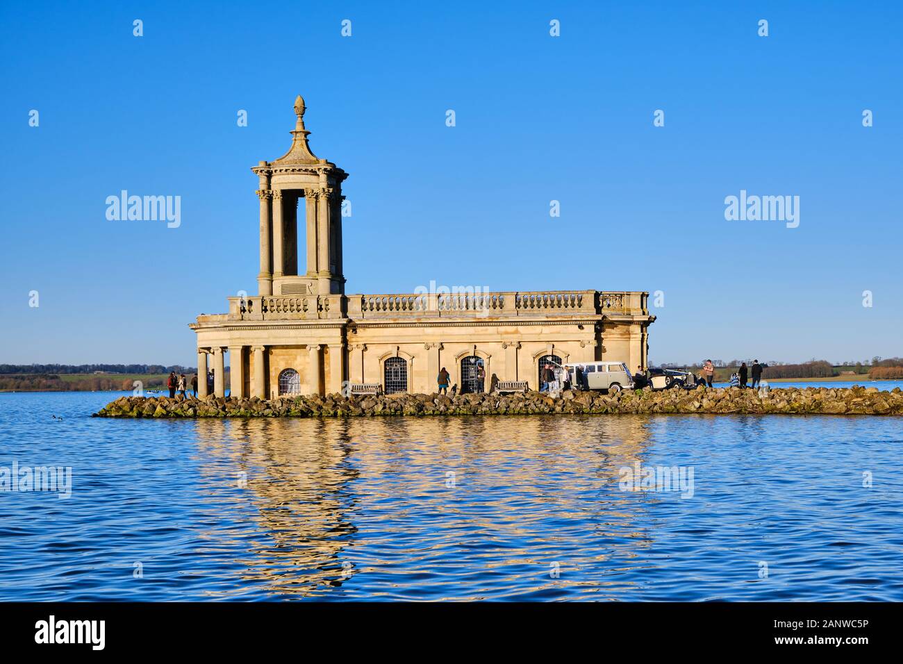Normanton Church situated on the Banks of Rutland Water with a cloudless vibrant blue sky behind and classic cars parked alongside Stock Photo