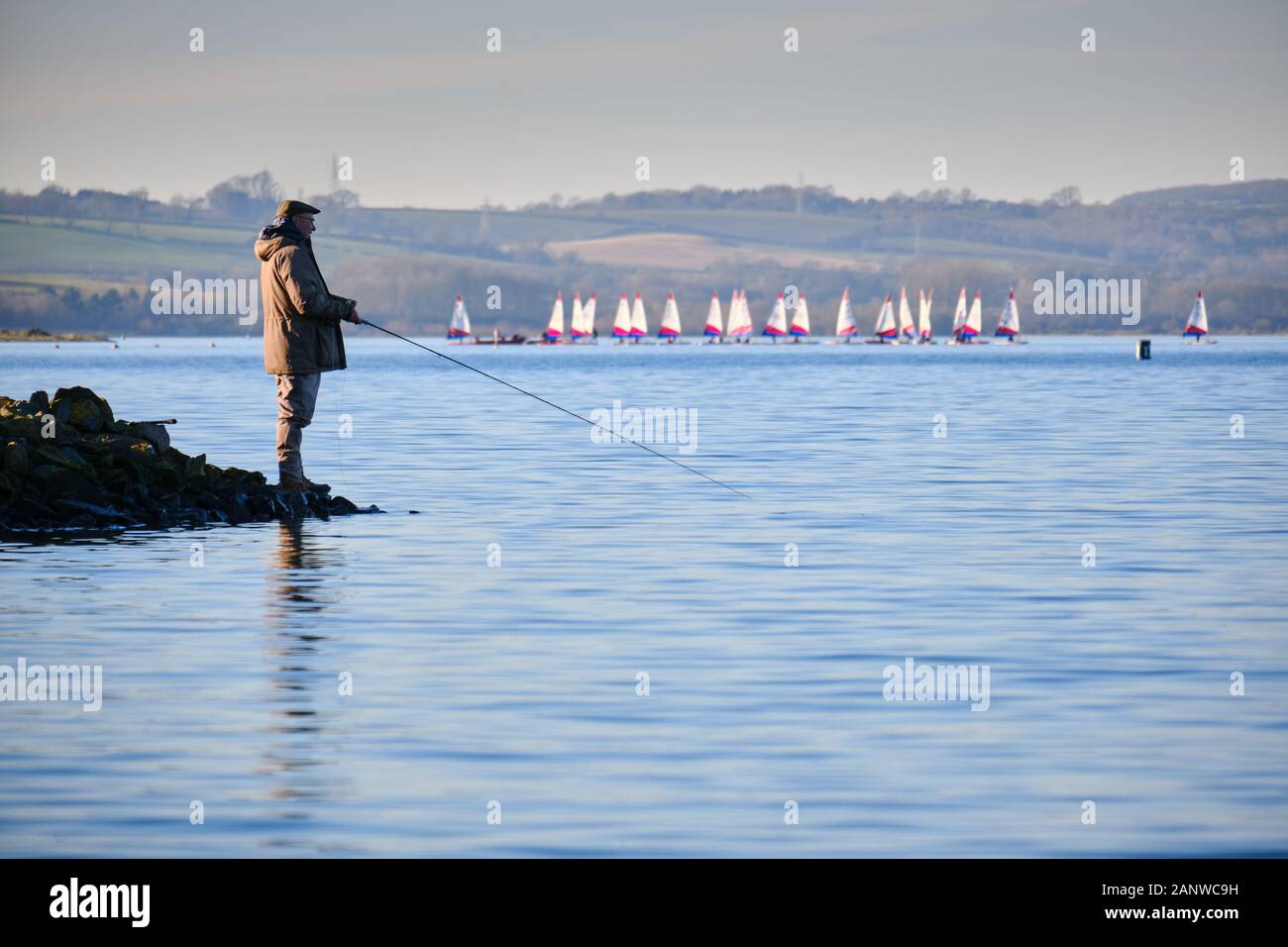 male angler fisherman sport fishing on the shore of Rutland Water an Anglian Water reservoir with a flotilla of small sailing boats in the background Stock Photo