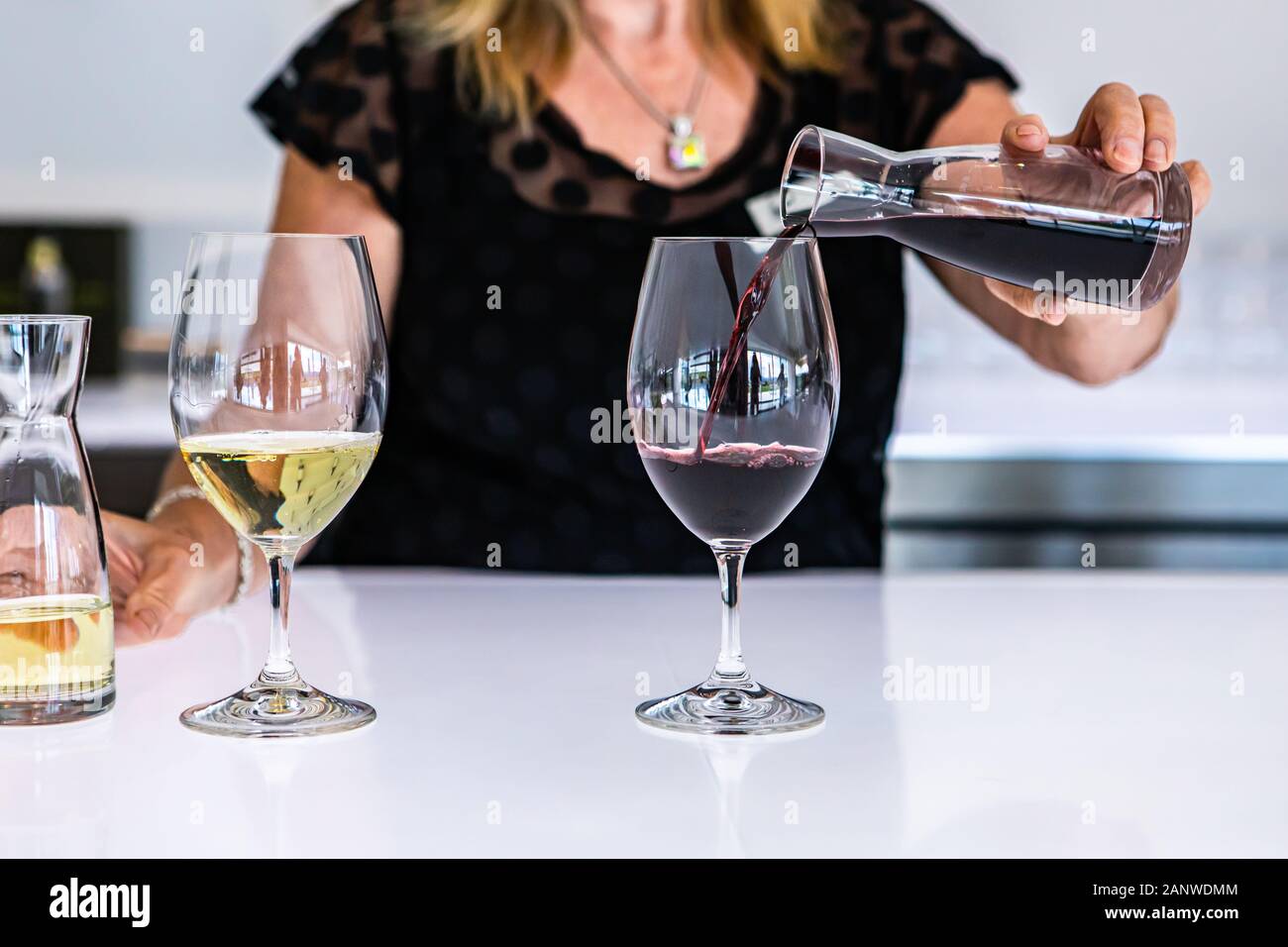 female bartender hand close up as she pouring red wine from a small mini decanter to the wineglass, next to a white glass of wine, bright bar counter Stock Photo