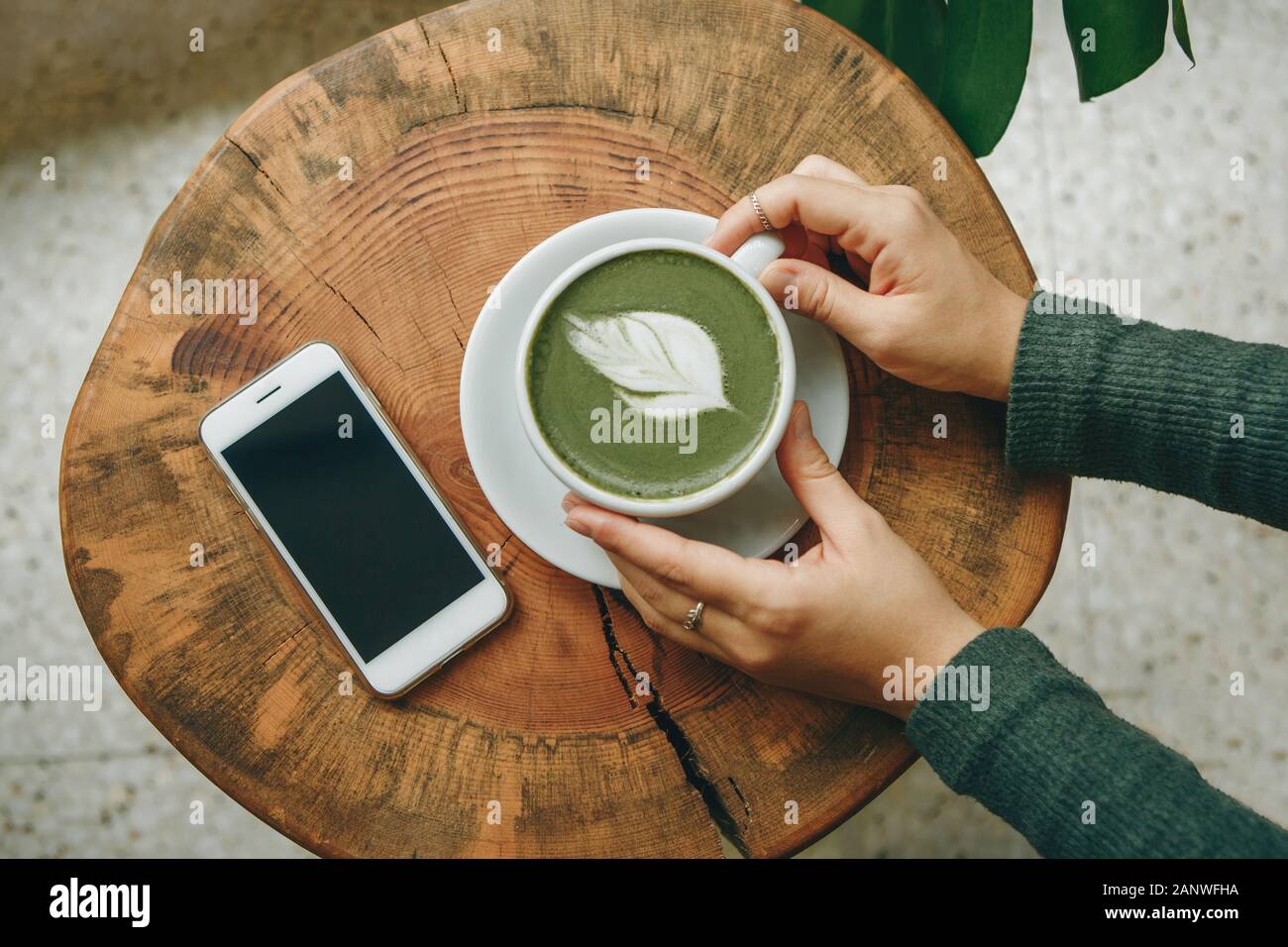 View from above. Girl holds in her hand a cup with fragrant fresh and healthy green matcha latte tea on a wooden table. Nearby lies a cell phone. Stock Photo