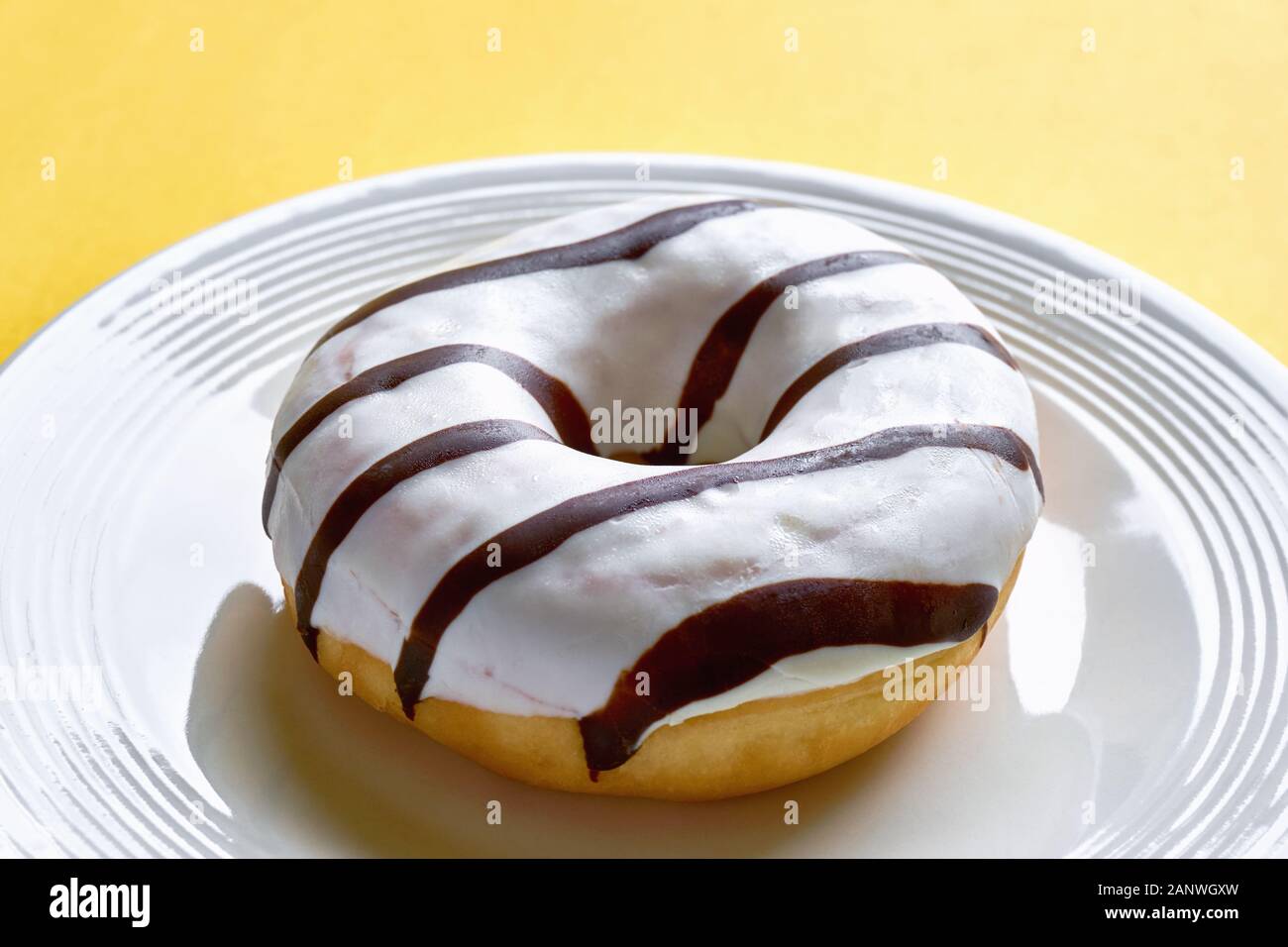 One single round donut with white ice glazing and stripes of dark brown chocolate on a shiny white plate against a yellow background Stock Photo