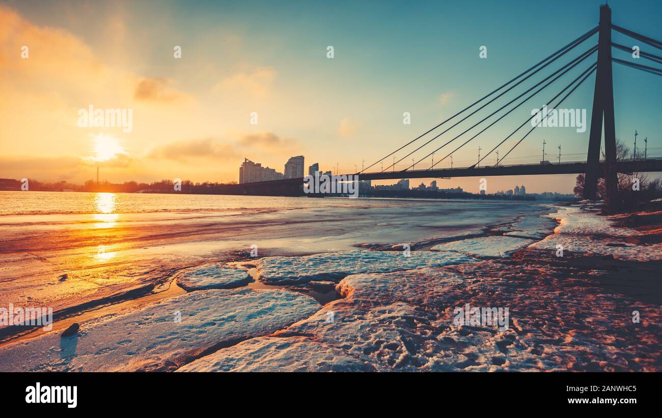 Pivnichnyi Bridge silhouette over half frozen Dnipro river against city at bright setting sun light. Concept river landscape, urban bridge structure, transport, environment. Stock Photo