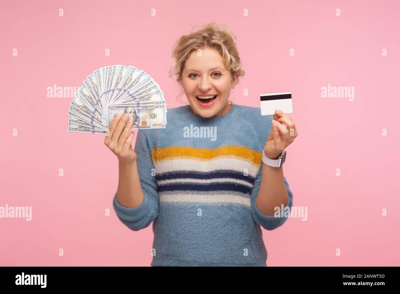 Portrait of surprised and happy adult woman with curly hair in sweater holding dollar banknotes and credit card, looking with shocked astonished expre Stock Photo
