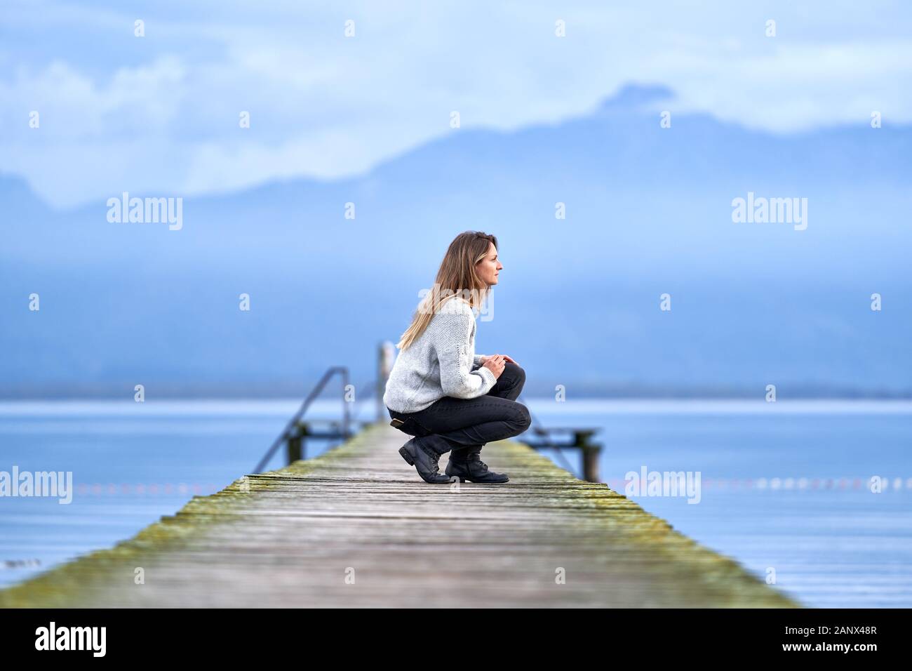 Young blond girl with a woolen pullover, squatting on a wooden pier in a calm lake of Bavaria. Picturesque composition with blue mountains and clouds Stock Photo
