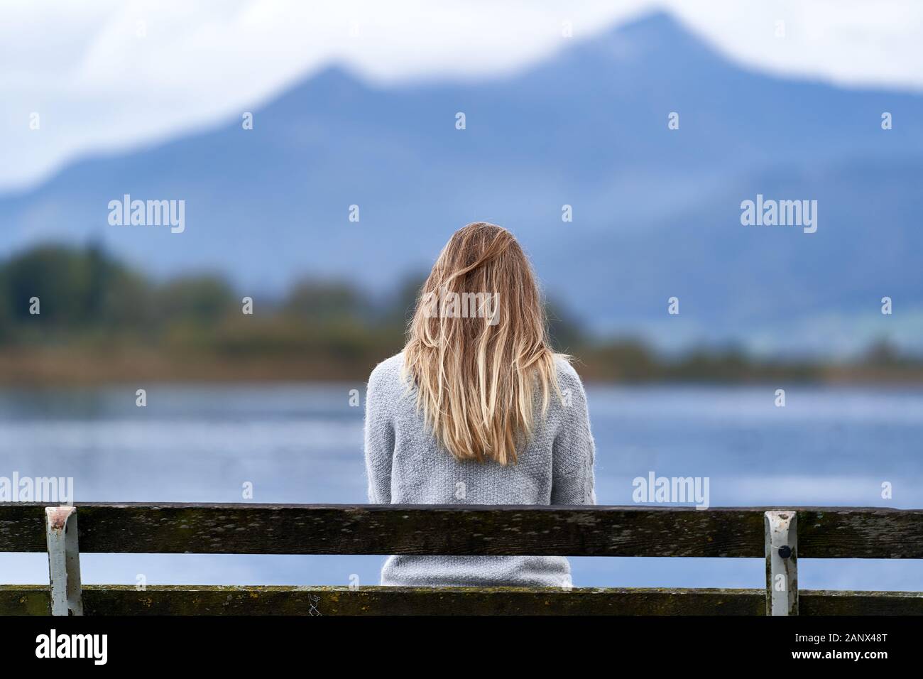 Young blond girl with long hair, sit down back to the camera on a wooden bench in front of a calm lake of Baviera. Picturesque composition with blue m Stock Photo