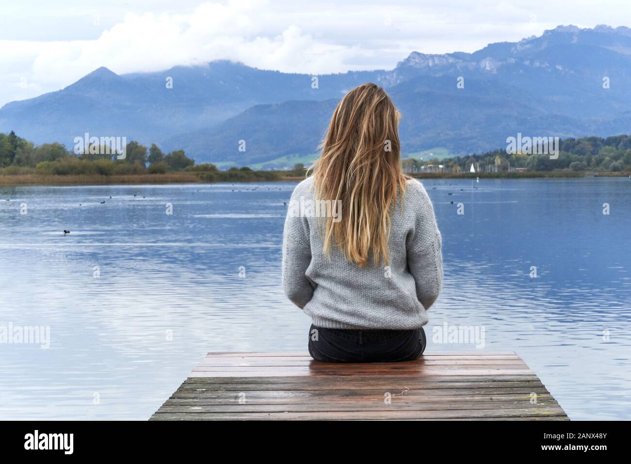 Young blond girl with long hair, sit down and back to the camera on a wooden pier in front of a calm lake looking to the horizon in Baviera. Travel co Stock Photo