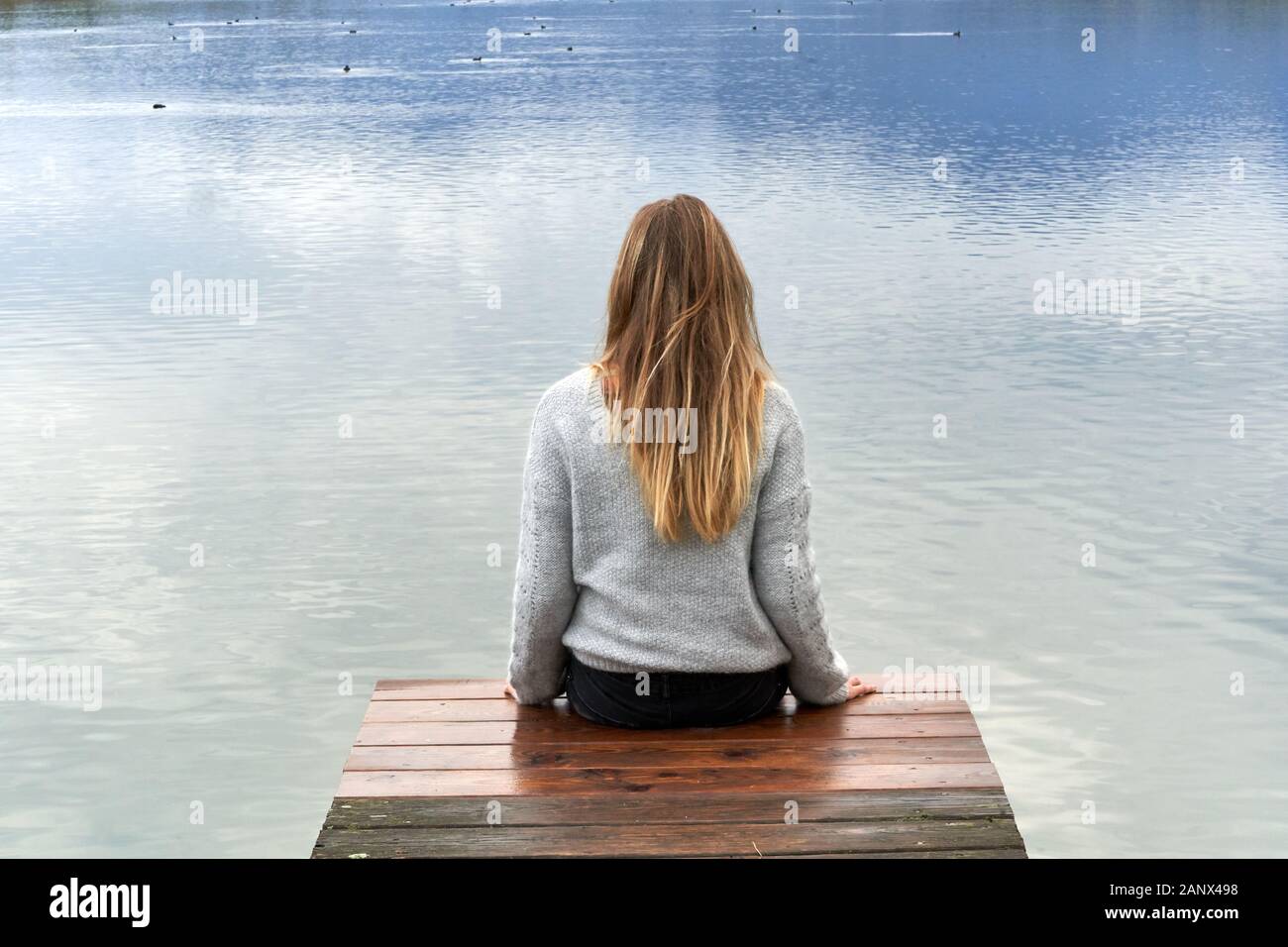 Young blond girl with long hair, sit down and back to the camera on a wooden pier in front of a calm lake looking to the horizon in Baviera. Travel co Stock Photo