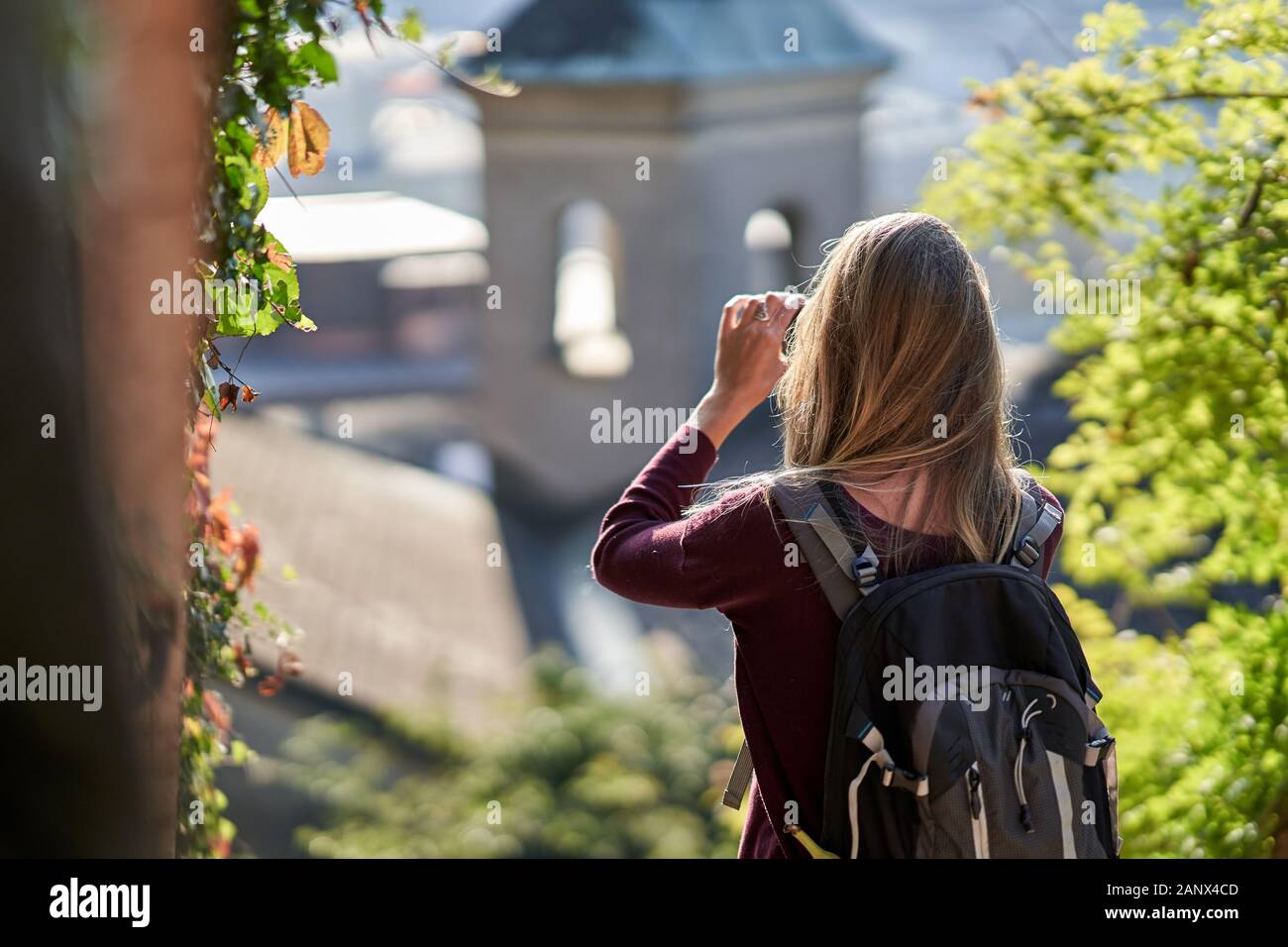 Young attractive woman standing backward with a backpack taking a picture of the old area of Salzburg with the sunset light. Travel concept. Stock Photo