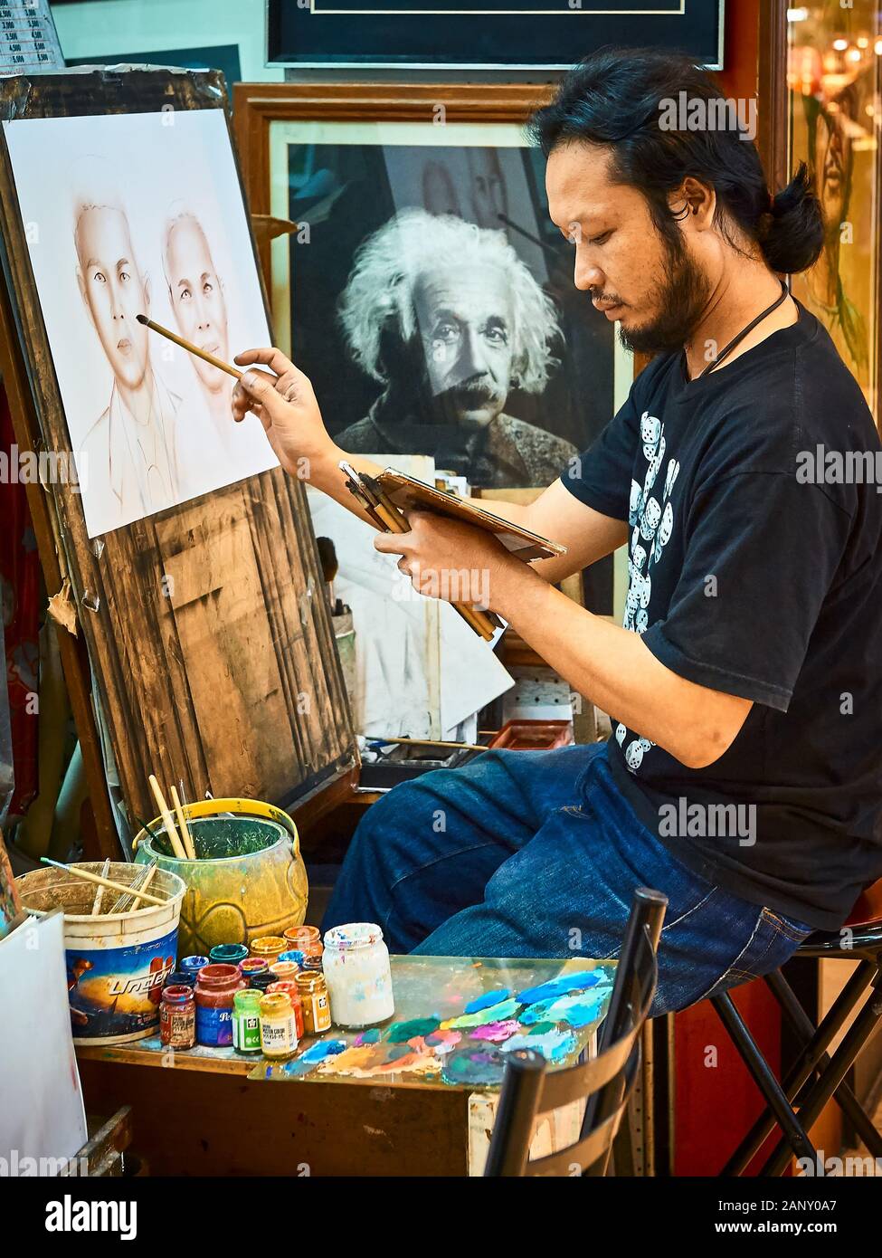 Bangkok, Thailand: Young man sits at a market painting a portrait of a couple; a picture of a watching Einstein hanging on the wall Stock Photo