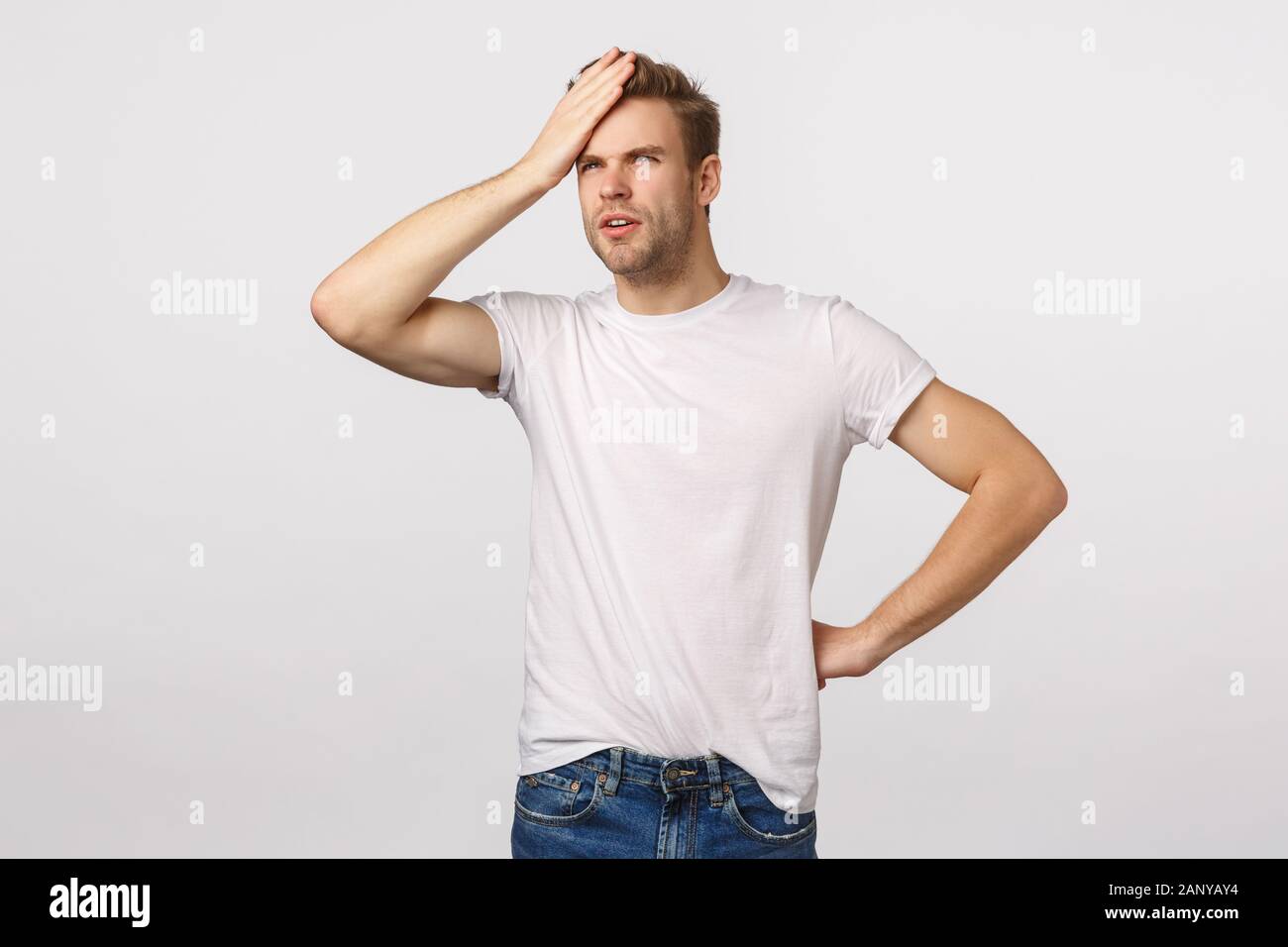 Focused and thoughtful young handsome blond bearded male in white t-shirt, punch forehead, squinting and looking upper left corner at sun, feeling Stock Photo