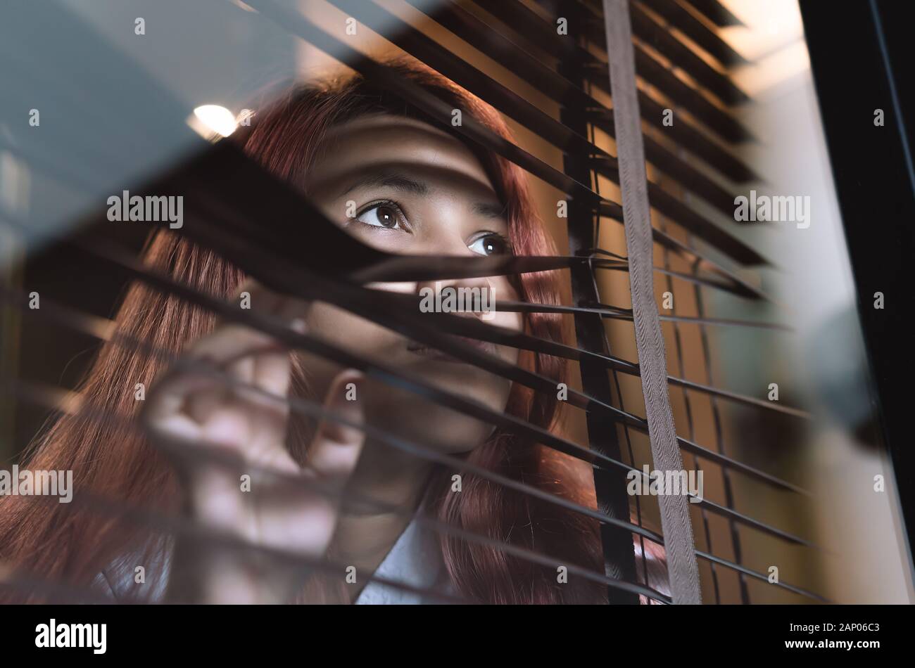 Asian woman looking through window blinds spying on neighbours - Young lonely millennial woman peeping through glass observing gossip and action outdoors - introvert, spy and intrusive concepts Stock Photo