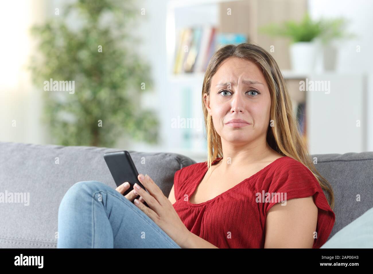 Frustrated woman holding smart phone looks at camera sitting on a couch ...