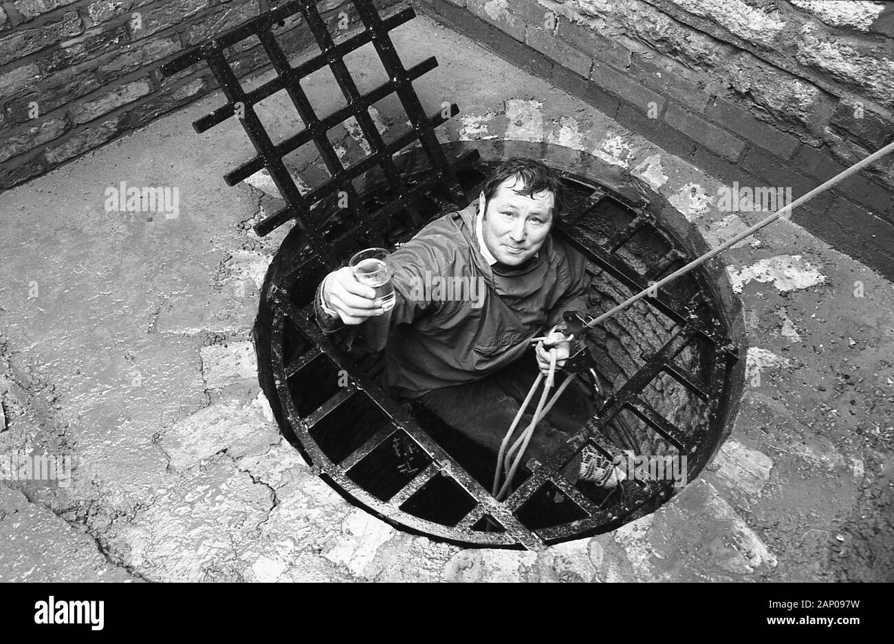 1980s, historical, a man holding onto a rope at the top of a well, showing a beaker of clean water he has got from the deep water well inside a church, England, UK Stock Photo