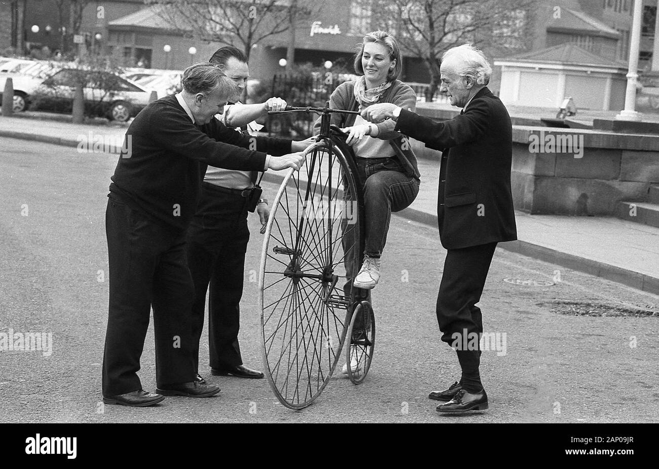 1980s, a young woman attempting to get on a penny-farthing bicycle, being held steady by some male helpers. Also known as the 'high wheeler' - as can be seen why in the picture - the machine was the first to be called a 'bicycle'. The 'ordinary' as it was also known as, had a large front wheel providing speed and comfort but became obsolete with the new 'safety' bicycles from the 1880s, which provided similar speed, comfort from their pneumatic tyres and with a reduced danger due far lower height to fall from. The name came from the British ''penny' and 'farthing' coins and symbolic of its era Stock Photo