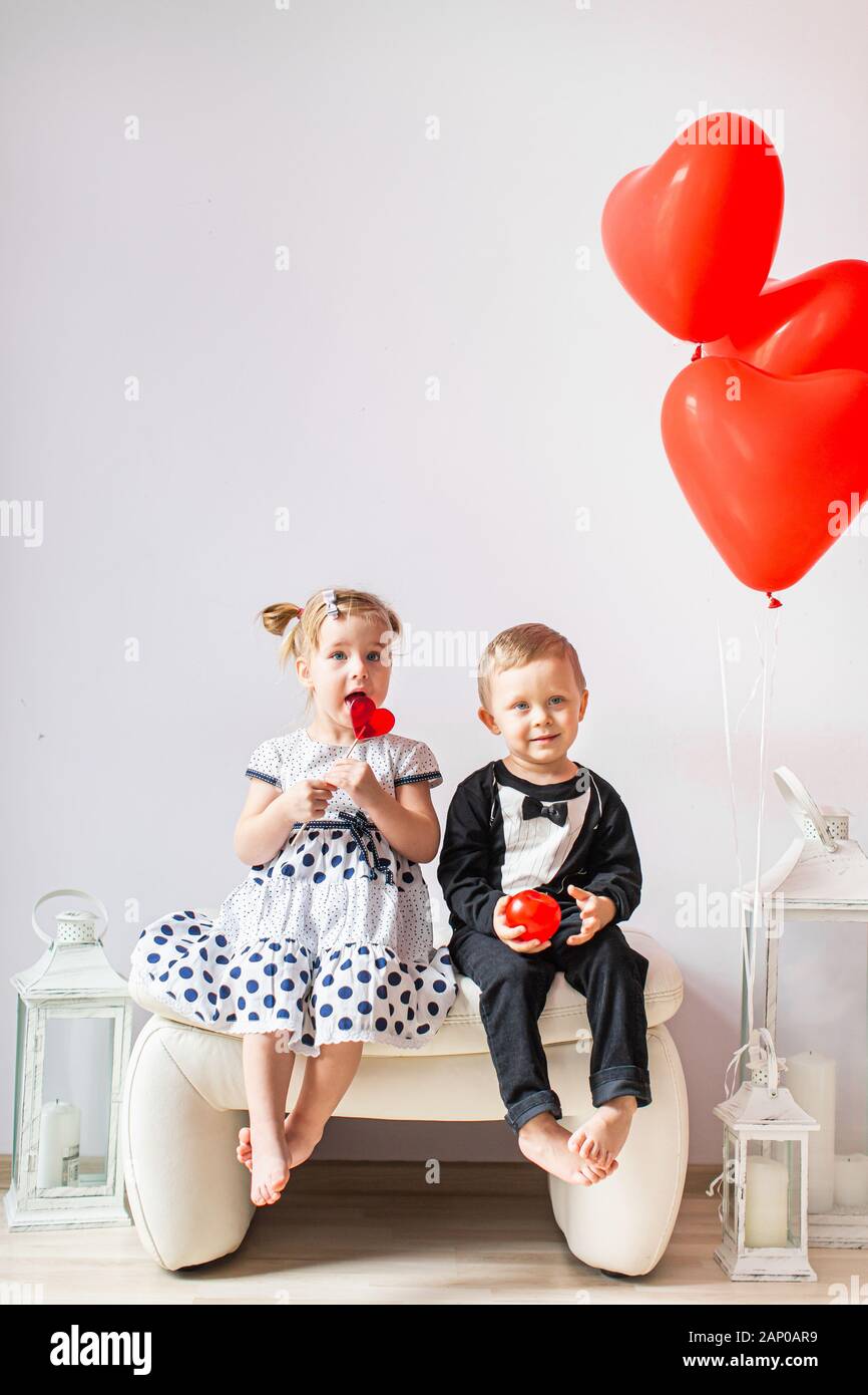 Little girl and boy sitting on a white chair near heart-shaped baloons. Girl licking a red lollipop. Valentines day concept Stock Photo