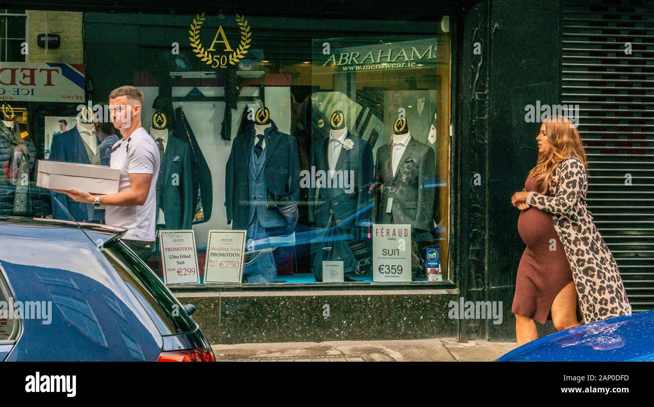 Man carrying box walking past shop selling suit and pregnant woman following behind in the St Stephen's Green area of Dublin in Ireland. Stock Photo