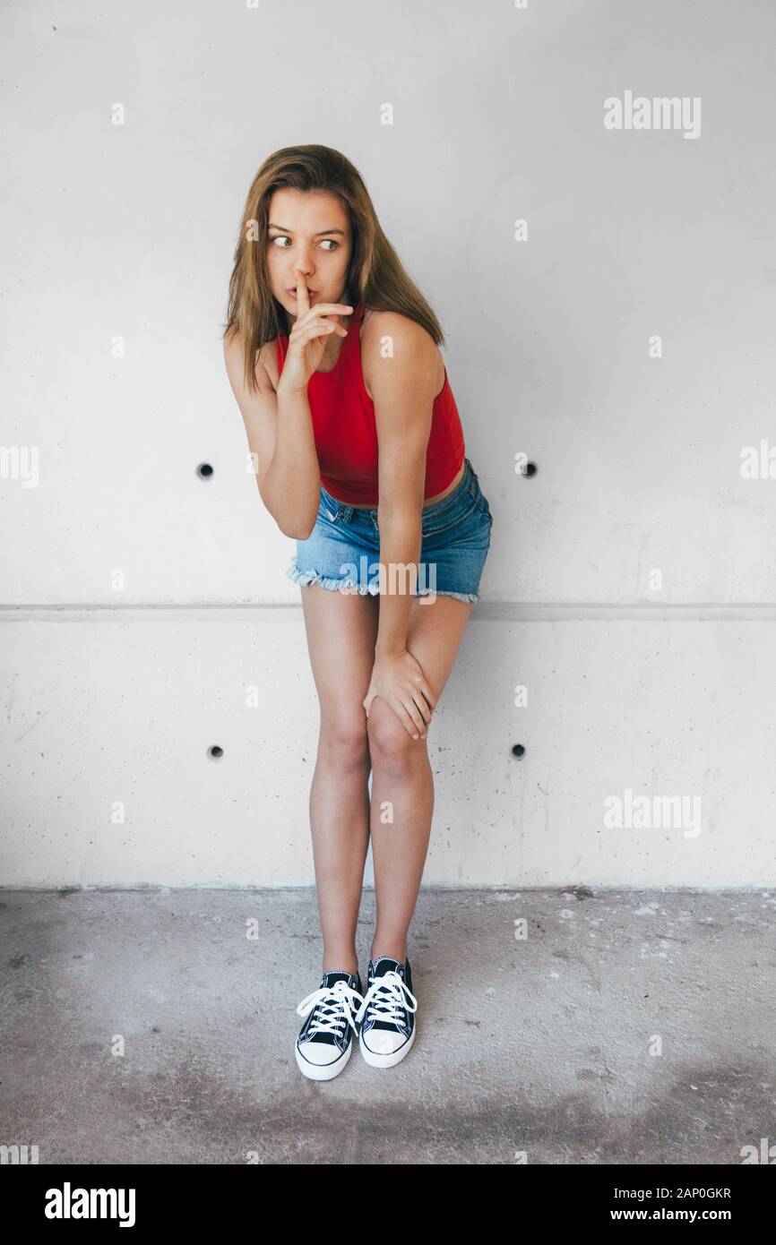 Teen girl in red top, denim mini skirt and sneakers doing silence sign with  finger and eyes wide opened against a concrete wall Stock Photo - Alamy