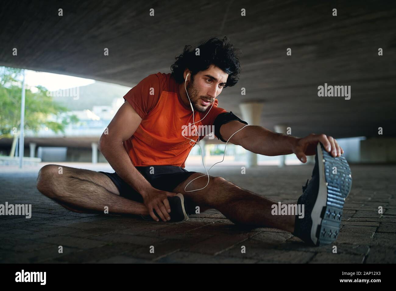 Young man athlete with earphone in his ears warming up and stretching his legs before running in the city street under the bridge Stock Photo