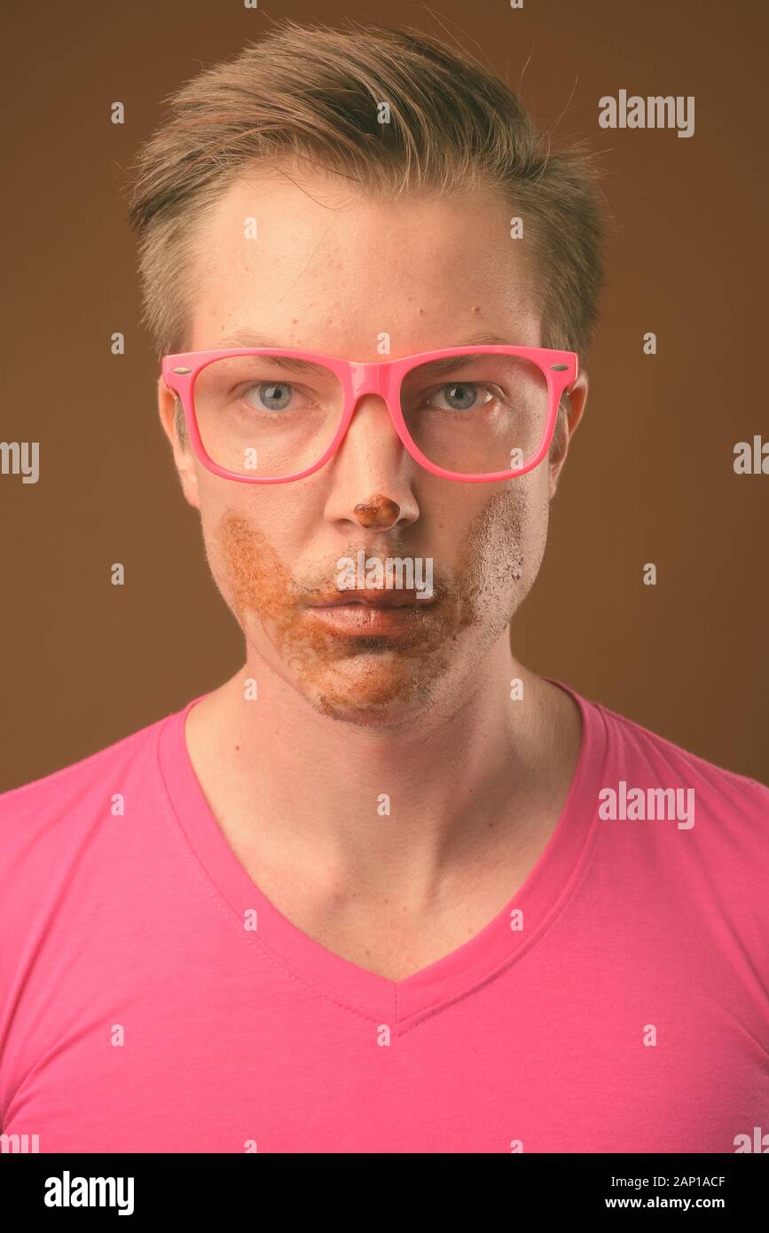 Studio shot of young handsome nerd man eating chocolate cake as messy concept against brown background Stock Photo