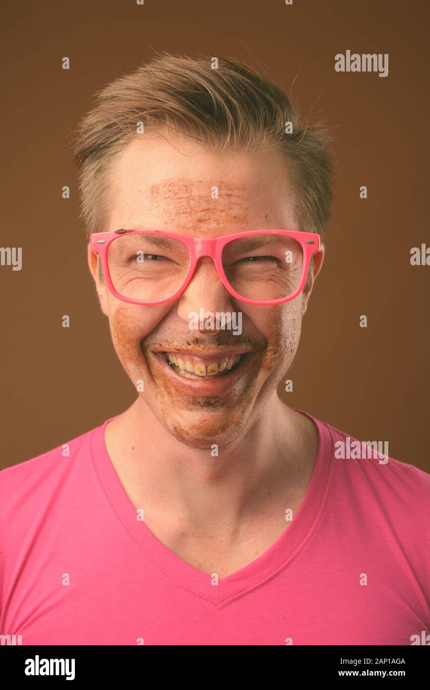 Studio shot of young handsome nerd man eating chocolate cake as messy concept against brown background Stock Photo